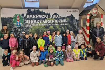 dzieci pozujące do zdjęcia wraz ze swoimi opiekunami w sali tradycji Karpackiego Oddziału Straży Granicznej. w tle baner informacyjny z napisem Karpacki Oddział Straży granicznej imienia 1 pułku strzelców podhalańskich. po prawej stronie budka wartownicza w kolorach biło czerwonych.