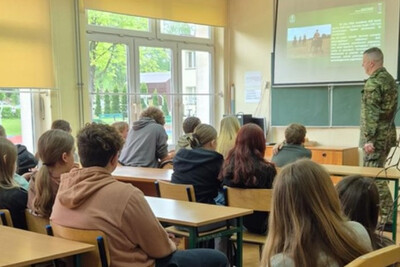 funkcjonariusz straży granicznej przemawiający do młodzieży szkolnej podczas prelekcji. wszystko odbywa się w klasie. na ścianie jest ekran z prezentacją omawianą przez funkcjonariusza.