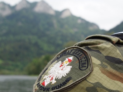 W tle trzy korony, na pierwszym planie ramię funkcjonariusza w umundurowaniu polowym. Zbliżenie na  emblemat z napisem straż graniczna polish border guard z orłem białym i słupkiem granicznym biało-czerwonym