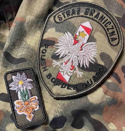 Zdobyli Wojskową Odznakę Górską 21 Brygady Strzelców Podhalańskich emblemat straży granicznej z napisem Straż Graniczna pod napisem znajduje się słupek graniczny w kolorze białoczerwonym a na na nim orzeł biały. pod słupkiem granicznym jest napis Polish Border Guard. obok emblematu straży granicznej znajduje się odznaka Górska na której wyszyta jest szarotka w kolorze białym z żółtym elementem, pod szarotką jest parzenica góralska w kolorach zielonym i żółtym. całość ułożona jest na mundurze straży granicznej typu polo.