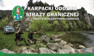 Na tle pienin stoi samochód służbowy SG i dwóch funkcjonariuszy. jeden z nich patrzy przez lornetkę, a drugi wskazuje palcem teren