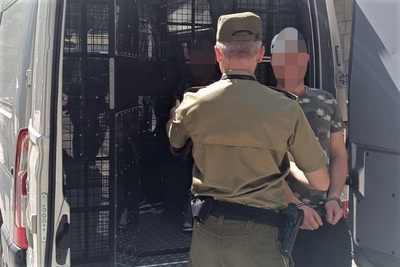 jeden z zatrzymanych cudzoziemców siedzi w samochodzie typu więźniarka a drugi jest prowadzony w kajdankach przez umundurowanego funkcjonariusza sg