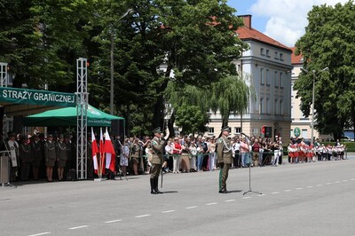 Komendant Karpackiego Oddziału Straży Granicznej wita wszystkie pododdziały zebrane na uroczystości. Za nim stoi i salutuje szablą dowódca uroczystości. W tle stoją wszyscy zebrani zaproszeni goście oraz rodziny funkcjonariuszy straży granicznej.