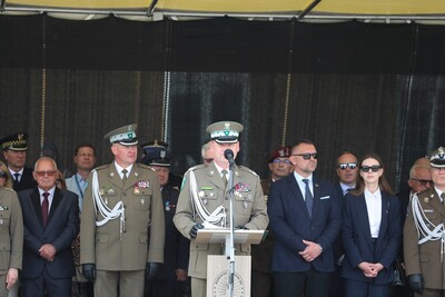 Komendant Karpackiego Oddziału Straży Granicznej przemawia do zebranych na uroczystości. Za nim stoją zaproszeni na uroczystość goście. Przed komendantem znajduje się podest z napisem Karpacki Oddział SG i z logiem oddziału a przed podestem znajduje się statyw w kolorze srebrnym na którym znajduje się mikrofon.