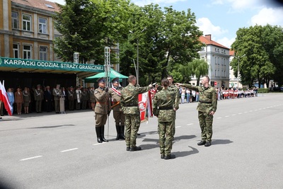 czworo nowo przyjętych funkcjonariuszy straży granicznej ubranych w mundury typu moro ślubują na sztandar Karpackiego oddziału straży granicznej. Stoją przed sztandarem z wyciągniętymi ku niemu prawymi rękami. Dwa palce w dłoni są skierowane ku sztandarowi. Sztandar trzyma funkcjonariusz straży granicznej. W tle jest trybuna gdzie stoją zaproszeni goście. Ślubowanie przyjmuje Komendant Karpackiego Oddziału Straży Granicznej. W tle obok trybuny stoją obecne rodziny funkcjonariuszy.