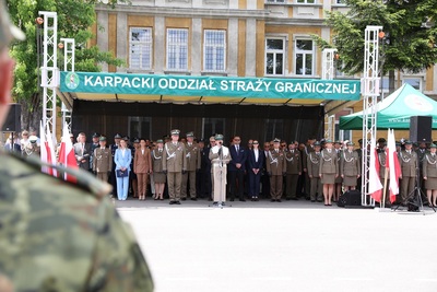 kapelan karpackiego oddziału straży granicznej błogosławi nowo przyjętym funkcjonariuszom. Przed nim stoi podest z napisem Karpacki Oddział SG i z logiem oddziału a przed podestem znajduje się statyw w kolorze srebrnym na którym znajduje się mikrofon. Za kapelanem znajdują się zaproszeni goście oraz Komendant Karpackiego Oddziału Straży Granicznej.