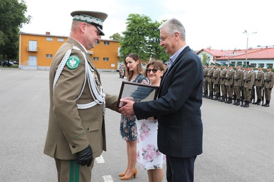 Komendant Karpackiego oddziału straży granicznej wręcza odznaczenia pracownikom karpackiego oddziału straży granicznej. W tle stoją pododdziały zebrane na uroczystości. Na dalszym planie widoczny jest budynek w kolorze pomarańczowym.