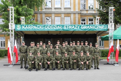 grupa nowo przyjętych funkcjonariuszy stoi w trzech szeregach, ustawiła się do zdjęcia grupowego. w tle trybuna oraz jeden z budynków komendy oddziału. po bokach powiewają biało-czerwone flagi.