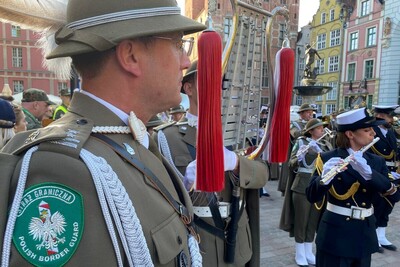naczelnik orkiestry reprezentacyjnej straży granicznej podczas wspólnego koncertu dwóch orkiestr straży granicznej. na pierwszym planie widoczny jest emblemat straży granicznej w kolorze zielonym na którym znajduje się kolejno od góry napis straż graniczna, poniżej jest słupek graniczny w kolorach białym i czerwonym oraz na nim orzeł biały w koronie , poniżej napis w języku angielskim polish border guard. w tle występują muzycy połączonych orkiestr straży granicznej. na dalszym planie widoczna jest fontanna z neptunem na rynku w Gdańsku.