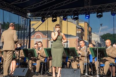 solistka orkiestry reprezentacyjnej straży granicznej wykonuje utwór.  w tle znajdują się muzycy orkiestry którzy koncert wykonują na siedząco. orkiestra dyryguje jej naczelnik.