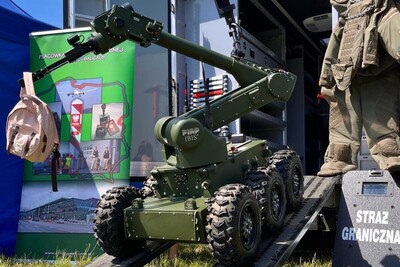 robot minerski zjeżdża z podestu. w chwytaku trzyma plecak w kolorze różowym. który wcześnie samodzielnie chwycił. w tle widoczny jest baner placówki w Krakowie Balicach. po prawej stronie zdjęcia widoczna jest tarcza ochronna z napisem straż graniczna a nad nią kombinezon saperski.