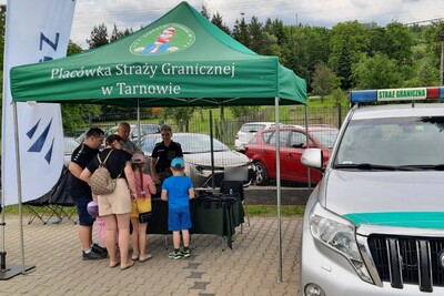 dzieci prawdopodobnie ze swoimi rodzicami zebrane przy stoisku straży granicznej. obok po prawej stronie zdjęcia widoczny jest pojazd służbowy straży granicznej.  stoisko znajduje się pod zadaszeniem, na którym jest napisane Placówka Straży Granicznej w Tarnowie. powyżej znajduje się logo karpackiego oddziału straży granicznej.