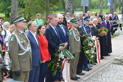zaproszeni goście zebrani podczas uroczystości. wśród nich przedstawiciele starostwa tatrzańskiego, urzędu miasta Zakopane oraz służb mundurowych. niektórzy w ręku trzymają wieńce lub kwiaty jak również znicze które zostaną złożone pod Pomnikiem.