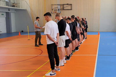 kandydaci stoją w szeregu w hali sportowej gdzie odbędzie się egzamin ze sprawności fizycznej. funkcjonariusz ubrany w strój wuefisty, omawia poszczególne ćwiczenia jakie kandydaci będą wykonywać. parkiet jest w kolorze pomarańczowo niebieskim z białymi liniami.