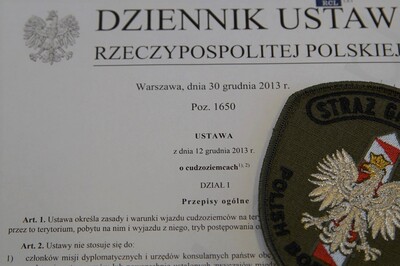 emblemat straży granicznej ułożony na papierowej wersji ustawy o cudzoziemcach. emblemat ma widoczny słupek graniczny w kolorze białoczerwonym oraz na nim orzeł biały. nad słupkiem znajduje się napis straż graniczna natomiast pod słupkiem polish border guard.