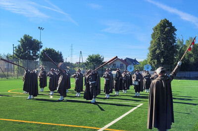 orkiestra reprezentacyjna straży granicznej podczas pokazu musztry paradnej. orkiestra dyryguje jej tamburmajor trzymający w ręku ciupagę zamiast buławy. orkiestr pokaz musztry paradnej wykonuje na boisku sportowym ze sztuczna murawą