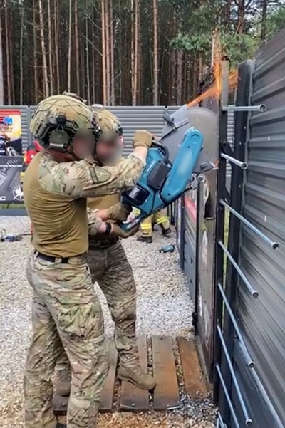 Dwóch funkcjonariuszy stoi przed metalowymi drzwiami. Jeden z nich trzyma w rękach piłę łańcuchową, która będzie otwierać siłowo metalowe drzwi.