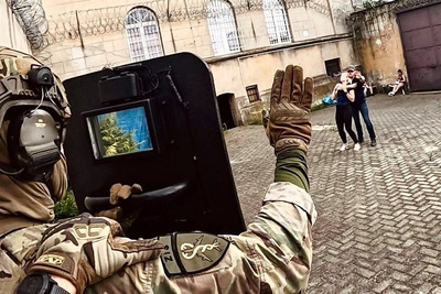 Dziedziniec więzienia. W tle jeden z mężczyzn trzyma jako zakładniczkę kobietę. Funkcjonariusz SG chroniąc się za tarczą ochronną trzyma wyciągniętą rękę, w geście &quot;spokojnie&quot;.