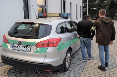 Z radiowozu SG wysiadł zatrzymany gruzin. jest prowadzony przez funkcjonariusza sg