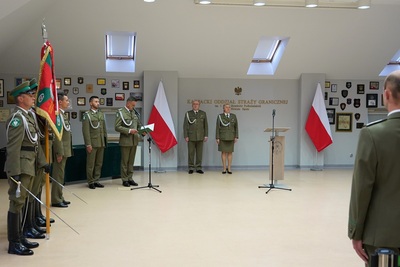 na zdjęciu widoczny poczet sztandarowy wraz ze sztandarem karpackiego oddziału straży granicznej. na drugim planie znajdują się po lewej stronie prowadzący uroczystość funkcjonariusze straży granicznej oraz na środku Pani komendant Karpackiego oddziału straży granicznej wraz z zastępca komendanta. w tle widoczne biało czerwone flagi Polski.