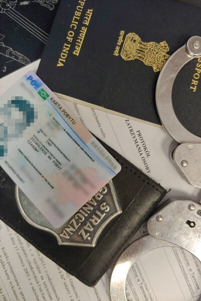 na zdjęciu widoczny jest paszport Indii w kolorze czarnym ze złotymi napisami oraz podrobiona polska karta pobytu. na wszystkim znajdują się kajdanki w kolorze srebrnym.