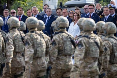 Żołnierze w hełmach defilują przed trybuną na której stoją przedstawiciele najwyższej władzy państwowej