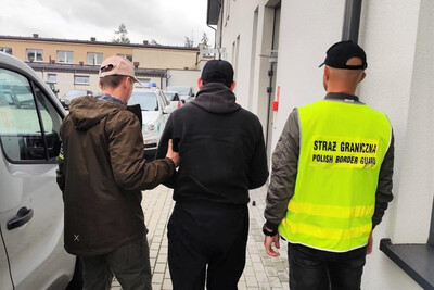 prowadzony przez funkcjonariuszy straży granicznej zatrzymany obywatel Kazachstanu. jeden z funkcjonariuszy ma na sobie założoną kamizelkę w kolorze żółtym z napisem straż graniczna a pod nim napis polish border guard w kolorze czarnym.