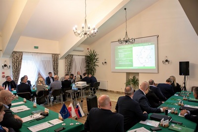 Uczestnicy spotkania siedzą przy stołach ułożonych w podkowę. Na ścianie na ekranie przedstawiana jest prezentacja dotycząca omawianych tematów.