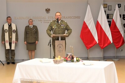 Komendant Karpackiego Oddziału Straży Granicznej przemawia do zabranych funkcjonariuszy oraz pracowników oddziału podczas spotkania opłatkowego. uroczystość odbyła się w sali tradycji oddziału. przed komendantem stoi stół przysłany w biały obrus z dekoracjami świątecznymi. za komendantem stoi jego zastępca oraz kapelan oddziału. widoczne są również flagi białoczerwone.