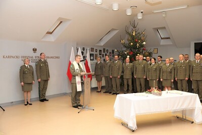 kapelan Karpackiego Oddziału Straży Granicznej przemawia do zabranych funkcjonariuszy oraz pracowników oddziału podczas spotkania opłatkowego. uroczystość odbyła się w sali tradycji oddziału. przed kapelanem stoi stół przysłany w biały obrus z dekoracjami świątecznymi. za kapelanem stoją komendanci oddziału oraz widoczne są białoczerwone flagi.