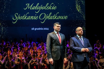 na zdjęciu widoczny wojewoda małopolski. przemawia do zebranych podczas małopolskiego spotkania wigilijnego. w tle widoczna duża grupa dzieci w strojach góralskich.