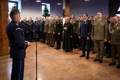 na pierwszym planie widoczny policjant który przemawia do wszystkich zebranych podczas spotkania opłatkowego. widoczne osoby równych służb mundurowych oraz księża.
