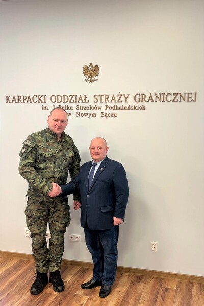 Komendant Karpackiego oddziału straży granicznej ściska dłoń funkcjonariusza który odchodzi na emeryturę. Pozują razem do zdjęcia w tle na ścianie widoczny jest napis w kolorze złotym Karpacki Oddział Straży Granicznej im. 1 Pułku Strzelców podhalańskich w  Nowym Sączu.