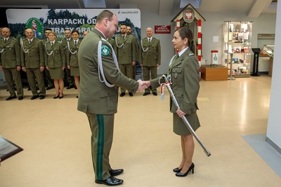 sala odpraw.  komendant kaosg podaje rękę i gratuluje odchodzącej na emeryturę funkcjonariuszce, która trzyma w rękach opuszczoną  szablę oficerską. w tle uczestnicy uroczystości