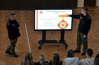 funkcjonariusze straży granicznej przeprowadzają prelekcje uczniom szkoły średniej. prelekcja odbywa się na sali gimnastycznej. przed funkcjonariuszami stoi duży ekran na którym wyświetlane są slajdy. uczniowie siedzą na krzesłach i słuchają prelekcji.