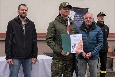 na zdjęciu widoczny funkcjonariusz straży granicznej który trzyma dyplom za zwycięstwo w biegu i ma na szyi zawieszony medal. obok stoi organizator zawodów. w tle widoczny budynek szkoły.
