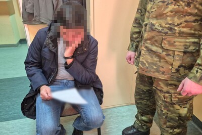 zatrzymany cudzoziemiec siedzi w pomieszczeniu służbowym straży granicznej. czyta swoje zeznania. obok niego widoczny funkcjonariusz straży granicznej który pilnuje zatrzymanego.