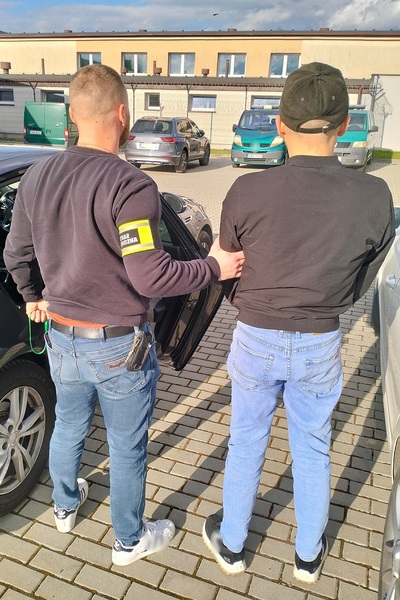 funkcjonariusz straży granicznej ubrany w cywilne ubrania trzyma za rękę zatrzymanego cudzoziemca. stoją na zewnątrz tyłem.