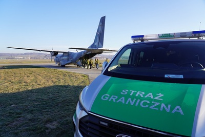 zbliżenie na przód samochodu służbowego sg typu bus.  w tle samolot wojskowy casa do którego wsiadają deportowani cudzoziemcy
