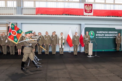 Hala sportowa. wchodzi poczet sztandarowy kaosg. w tle komendant i kadra kierownicza kaosg. na balustradzie nad nimi flaga i godło państwowe