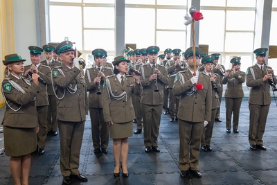 hala sportowa. orkiestra reprezentacyjna straży granicznej w pozycji stojącej gra na instrumentach, kieruje nią tamburmajor, dwie solistki i solista śpiewają hymn Polski