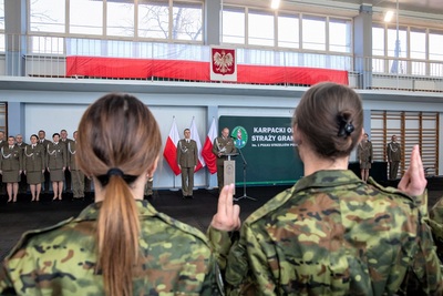 ujęcie z tyłu, dwie ślubujące funkcjonariuszki stoją z podniesionymi dłońmi z wysuniętymi palcami powtarzając rotę ślubowania. w tle komendant czytający rotę ślubowania, kadra kierownicza i prowadzący uroczystość. Na balustradzie nad nimi flaga i godło Polski