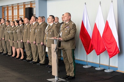 generał jopek stoi przy mównicy. przemawia do mikrofonu. obok niego w dwuszeregu stoi kadra kierownicza oddziału. za generałem trzy atłasowe biało-czerwone flagi na stajakach