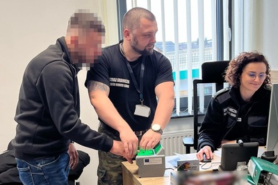Przy biurku siedzi funkcjonariuszka SG pracująca na komputerze. Funkcjonariusz stojący obok pobiera odciski palców na specjalnym urządzeniu od zatrzymanego cudzoziemca.