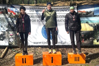 Na zdjęciu widoczna jest trójka uczestników w strojach sportowych, stoją na oznaczonym numerami podium, które jest koloru pomarańczowego.