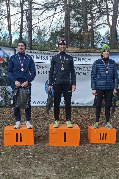 Na zdjęciu widoczna jest trójka uczestników w strojach sportowych, stoją na oznaczonym numerami podium, które jest koloru pomarańczowego.