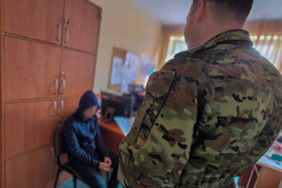 W pomieszczeniach służbowych straży Granicznej, funkcjonariusz Straży Granicznej ubrany w mundur stoi zwrócony do zatrzymanego i patrzy na niego. Zatrzymany siedzi na krześle ze wzrokiem zwróconym w podłogę.