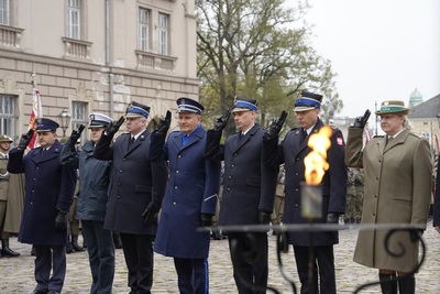 Na zdjęciu widać siedmiu funkcjonariuszy w mundurach różnych formacji, którzy oddają honor. W tle po lewej stronie widać budynek. Na pierwszym planie płonie ogień w świeczniku.