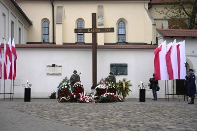 Na tle budynków widać duży krzyż z napisem Katyń 1940 -1990. Dwóch mężczyzn w mundurze z bronią stoją po obu stronach krzyża. Widać biało-czerwone kwiaty pod krzyżem oraz zapalone znicze. Po obu stronach zdjęcia widać flagi biało czerwone.