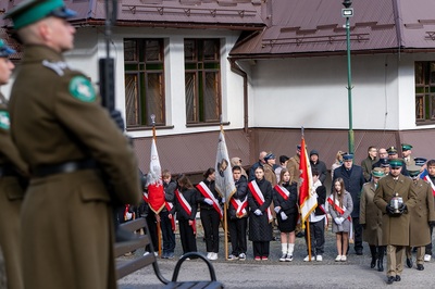 Na tle dużego białego budynku stoją dzieci z pocztami sztandarowymi. Osoby w mundurze idą ze zniczem. Po lewej stronie widać mężczyznę w mundurze.